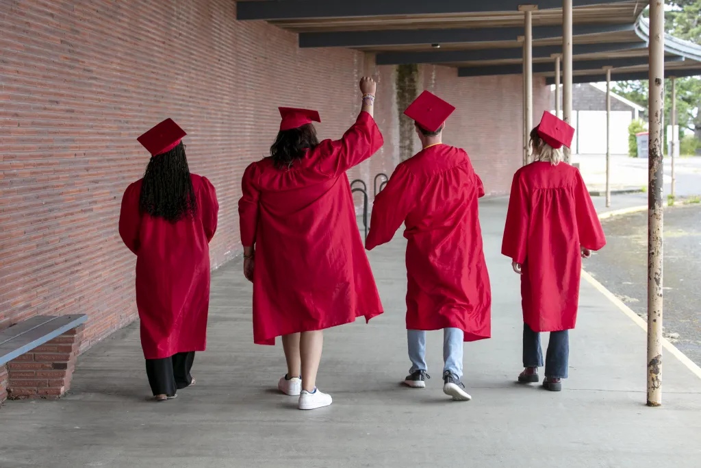 Four graduates in red caps and gowns walking away from the camera down a covered walkway, one with a fist raised in celebration.